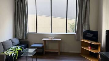 A living room with a sofa, a glass coffee table, a TV on a wooden stand, a small table with a white object, and a window with curtains.
