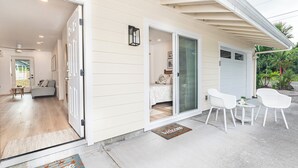 Une maison dotée d’une porte-fenêtre coulissante qui donne sur un espace de vie comprenant un canapé et une table basse. Il y a une petite terrasse avec deux chaises blanches et une table.