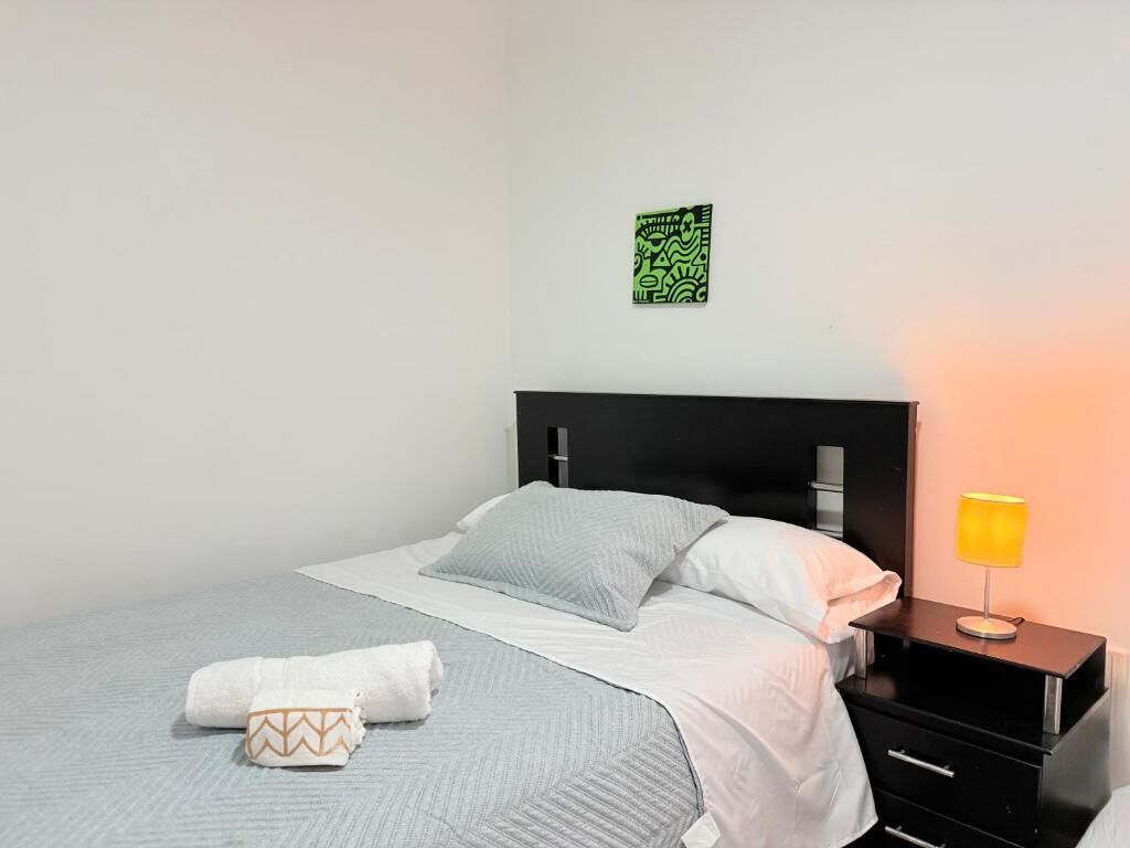 Una cama bien hecha con colcha gris y blanca, una almohada blanca y una toalla doblada en el suelo. Hay una mesita de noche negra con una lámpara amarilla al lado de la cama. Un cuadro abstracto verde cuelga de la pared sobre la cama.