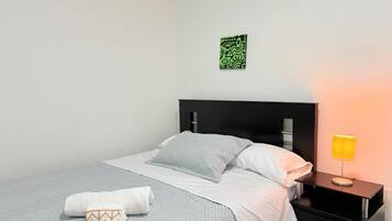 Una cama bien hecha con colcha gris y blanca, una almohada blanca y una toalla doblada en el suelo. Hay una mesita de noche negra con una lámpara amarilla al lado de la cama. Un cuadro abstracto verde cuelga de la pared sobre la cama.