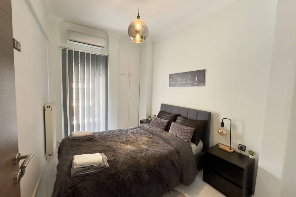 Ein Schlafzimmer mit einem großen Bett, einem Nachttisch mit Lampe, einem Wandbild und einem Fenster mit Jalousien.