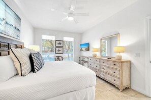 A bedroom with a large bed, a dresser, a ceiling fan, and a view of the outdoors. - Coastal Calm, Private Deeded Beach Access, 4 Seater Golf Cart, Bikes, Dogs (Inlet beach)