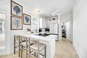 A modern kitchen with white cabinetry, a central island with bar stools, and framed artwork on the wall. - Coastal Calm, Private Deeded Beach Access, 4 Seater Golf Cart, Bikes, Dogs (Inlet beach)