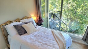 Une chambre avec un grand lit, une table de chevet, une lampe et une vue sur un balcon bordé d’arbres.
