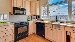 Une cuisine avec des meubles en bois, un poêle noir, un lave-vaisselle et une grande fenêtre donnant sur les arbres.