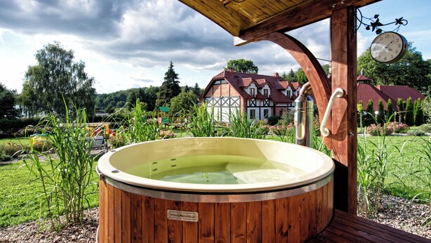 A wooden hot tub with a wooden deck, surrounded by greenery, with a traditional house in the background.