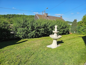 Ein Garten mit einem Steinfbrunnen, üppigem Grün und einem Haus im Hintergrund.