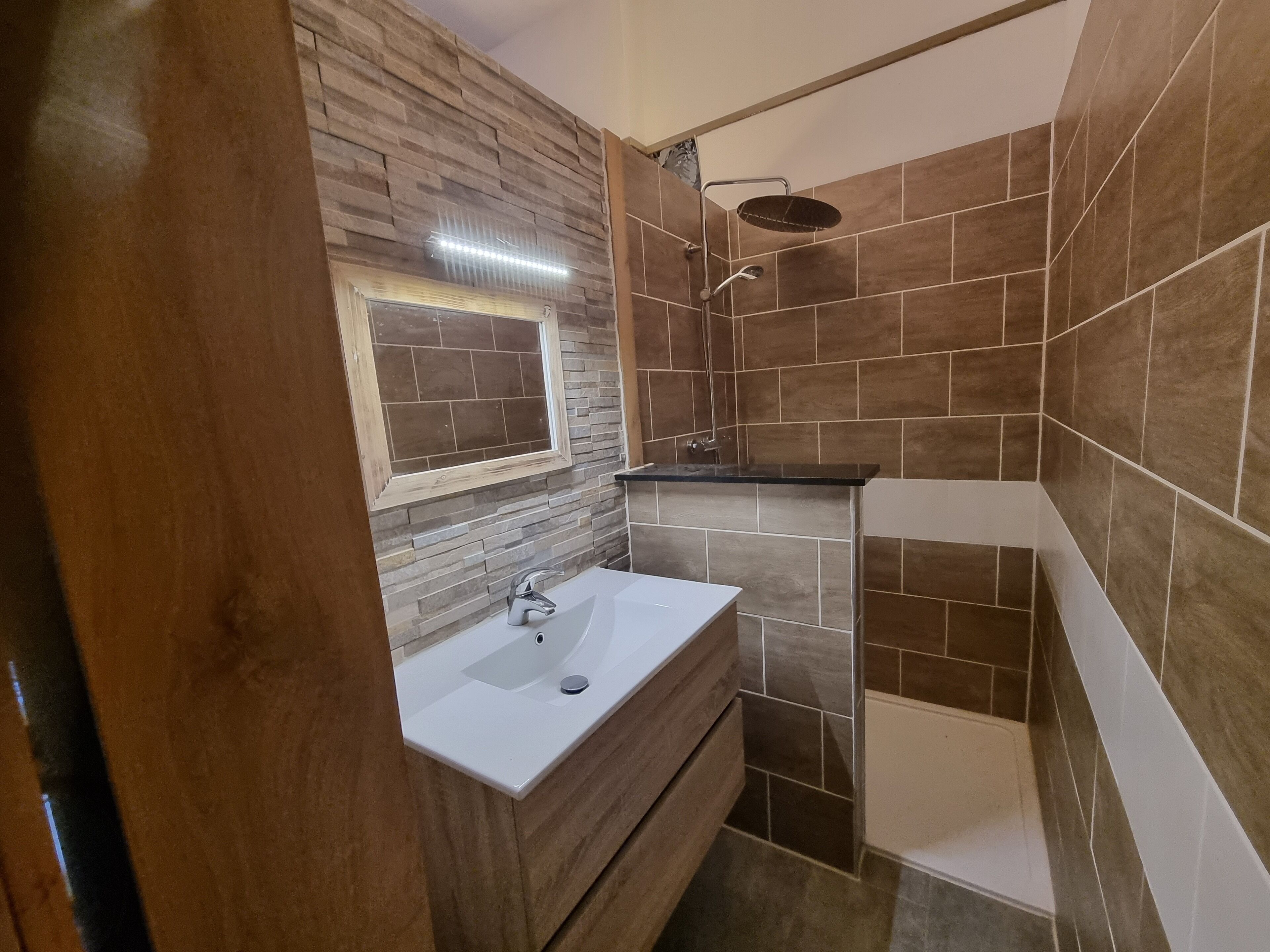 A bathroom with a white sink, a wooden vanity, a shower with a black countertop, and a window with a wooden frame.