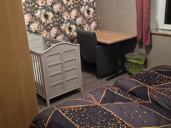 Ein Zimmer mit Reisebett, Schreibtisch, Stuhl, Mülleimer und einem Bett mit gemustertem Überwurf.