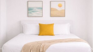 Una cama con ropa blanca, una almohada amarilla y un plaid color beige.