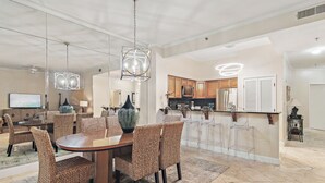 A spacious dining area with a round table and wicker chairs, a kitchen with wooden cabinets, and a bar area with stools. - 2BD/2BTH Penthouse - Beachfront BLDG Panoramic View - Tides 1511 (Miramar Beach)