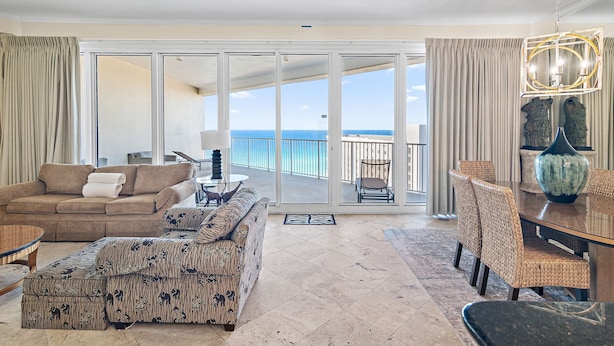 A spacious living area with a large sliding glass door leading to a balcony with a view of the ocean. There is a brown sofa, patterned ottoman, and a dining table with chairs. - 2BD/2BTH Penthouse - Beachfront BLDG Panoramic View - Tides 1511 (Miramar Beach)