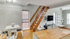 Uma sala de estar moderna com escada de madeira, área de jantar com cadeiras brancas, TV fixada na parede e mesa de centro de madeira.