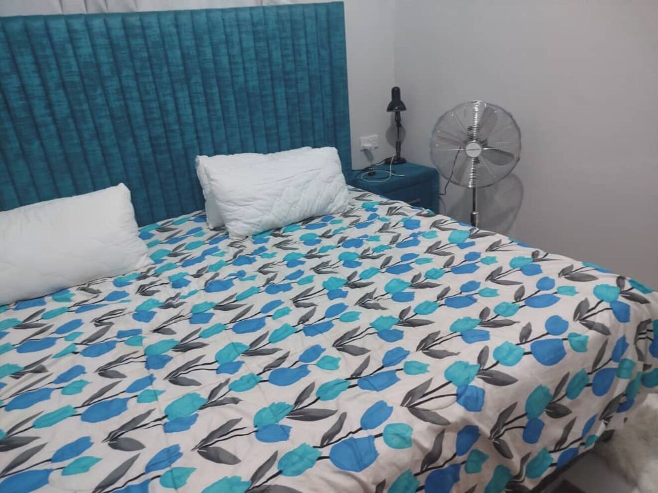 A bed with a patterned bedspread, a blue headboard, a bedside table with a lamp, and a standing fan.