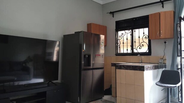 A compact living space with a flat-screen TV, a stainless steel refrigerator, a small kitchen area with a sink and a chair, and a window with decorative bars. - Quaint 1-bedroom apartment in pleasant Kampala neighborhood (Kampala)