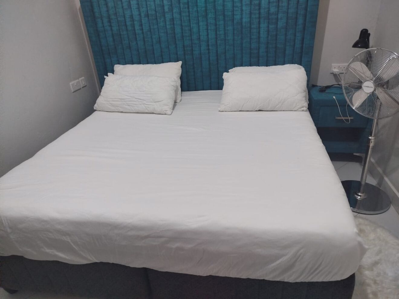Camera con un letto singolo, copripiumino bianco, due cuscini, tenda blu e ventola da terra.