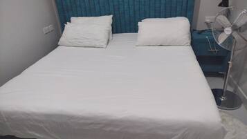 Camera con un letto singolo, copripiumino bianco, due cuscini, tenda blu e ventola da terra.