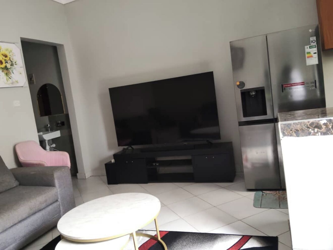 A modern living room with a flat-screen TV, a grey sofa, a small round table, and a kitchen area with a refrigerator.