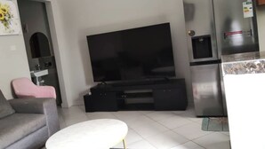 A modern living room with a flat-screen TV, a grey sofa, a small round table, and a kitchen area with a refrigerator. - Quaint 1-bedroom apartment in pleasant Kampala neighborhood (Kampala)