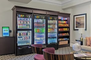 Eine Hotelhalle mit drei Getränkeautomaten, einer Sitzecke mit einem runden Tisch und einem Bücherregal mit Snacks.