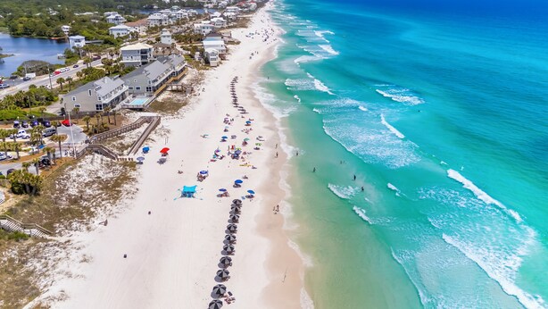 An aerial view of a beach with rows of beach umbrellas and people, a clear blue ocean, and buildings along the shore. - 30A Home for 10: Pool, Golf Cart & Chef's Kitchen, Open-Concept Living (Santa Rosa Beach)