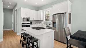 A modern kitchen with white cabinets, stainless steel appliances, a central island with stools, and a dining area with a table and chairs. - 30A Home for 10: Pool, Golf Cart & Chef's Kitchen, Open-Concept Living (Santa Rosa Beach)