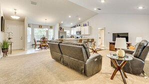 Una sala de estar moderna con un sofá, una mesa de centro y un televisor. Se observa una cocina al fondo con armarios blancos y un microondas.