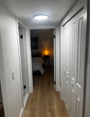 A hallway with wooden flooring leading to a bedroom with a bed and a bedside table with a lamp.