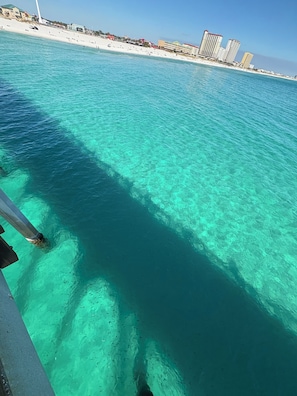 A clear turquoise sea with a wooden pier extending into it. In the background, there are buildings and a sandy beach. - Casa Blanca On The Canal (Gulf Breeze)