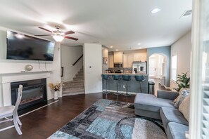 A modern living room with a fireplace, a large flat-screen TV, a ceiling fan, and a kitchen area with bar stools. - 3-bedroom Modern Townhome in charming Lithonia! (Lithonia)