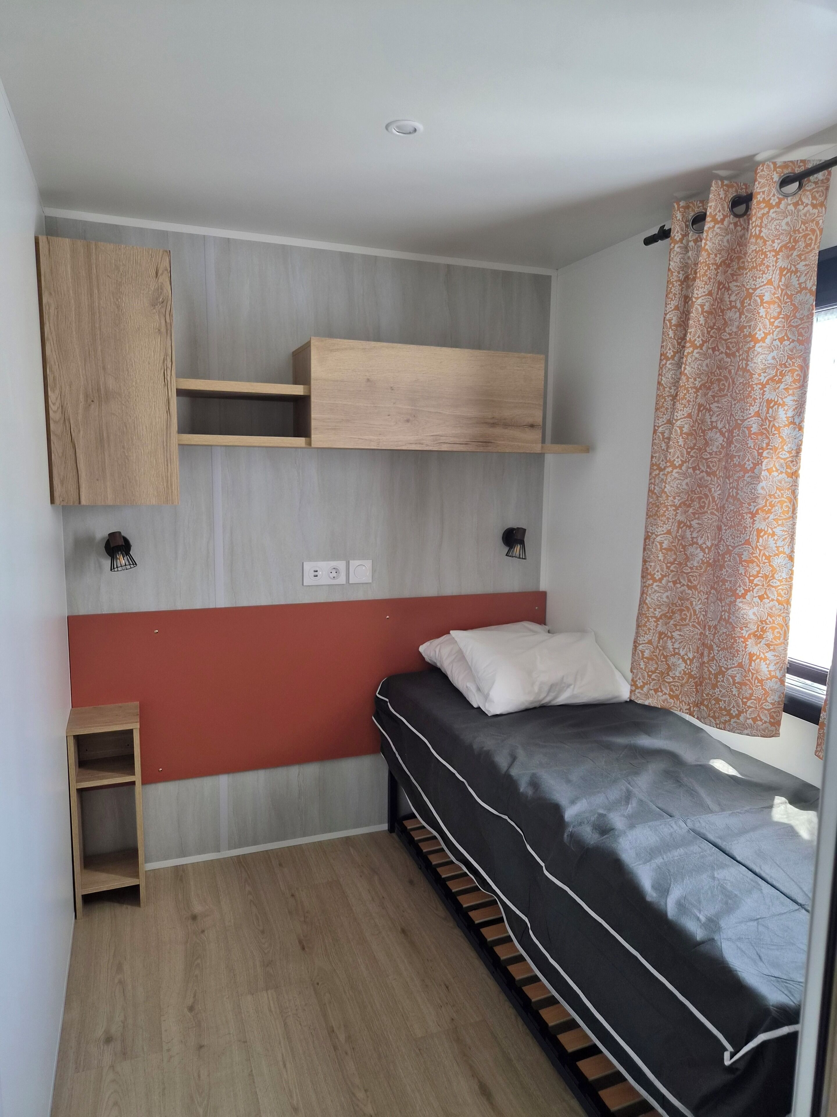 A small, compact room with a bed, a wooden shelf unit, a small table, and a window with curtains.