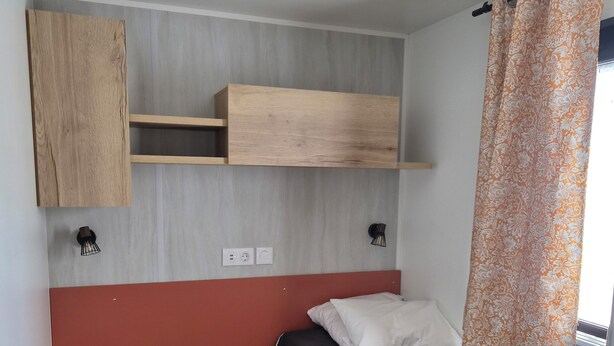 Ein kleiner, kompakter Raum mit einem Bett, einer Holzregaleinheit, einem kleinen Tisch und einem Fenster mit Vorhängen.