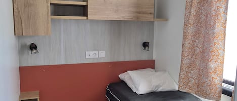 A small, compact room with a bed, a wooden shelf unit, a small table, and a window with curtains.