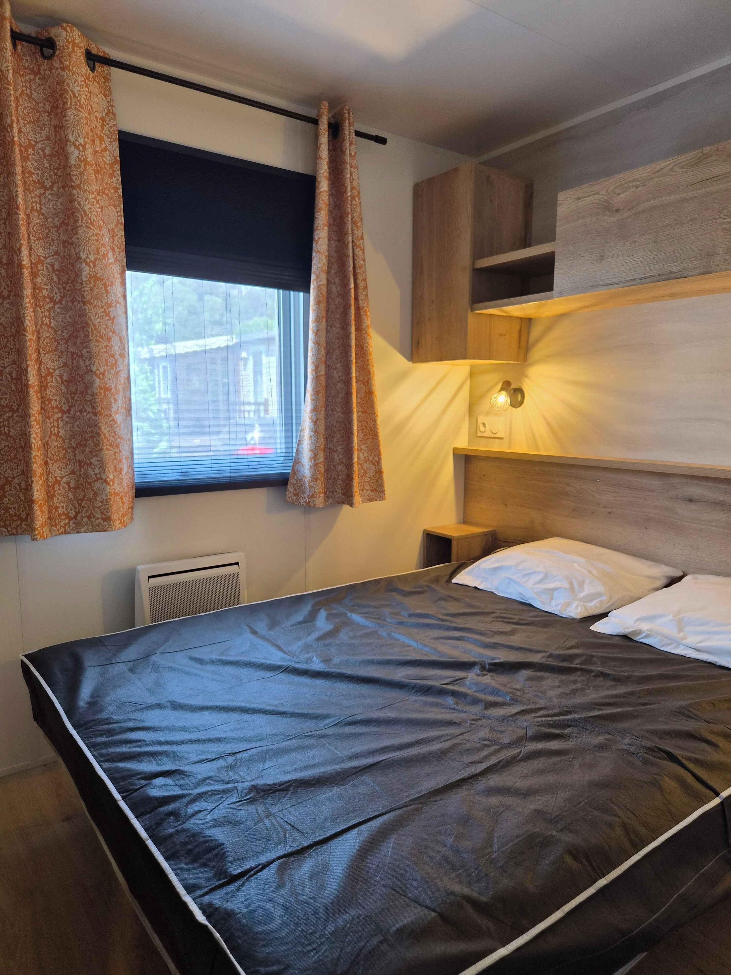 A bed with a dark mattress, wooden headboard, and white pillows. There is a window with curtains, a wall-mounted light fixture, and a small bedside table.