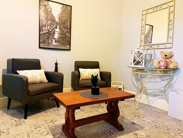 Un salón con dos sillones oscuros, una mesa de centro de madera, una mesita auxiliar con un peluche, un cuadro enmarcado en la pared y un espejo decorativo.