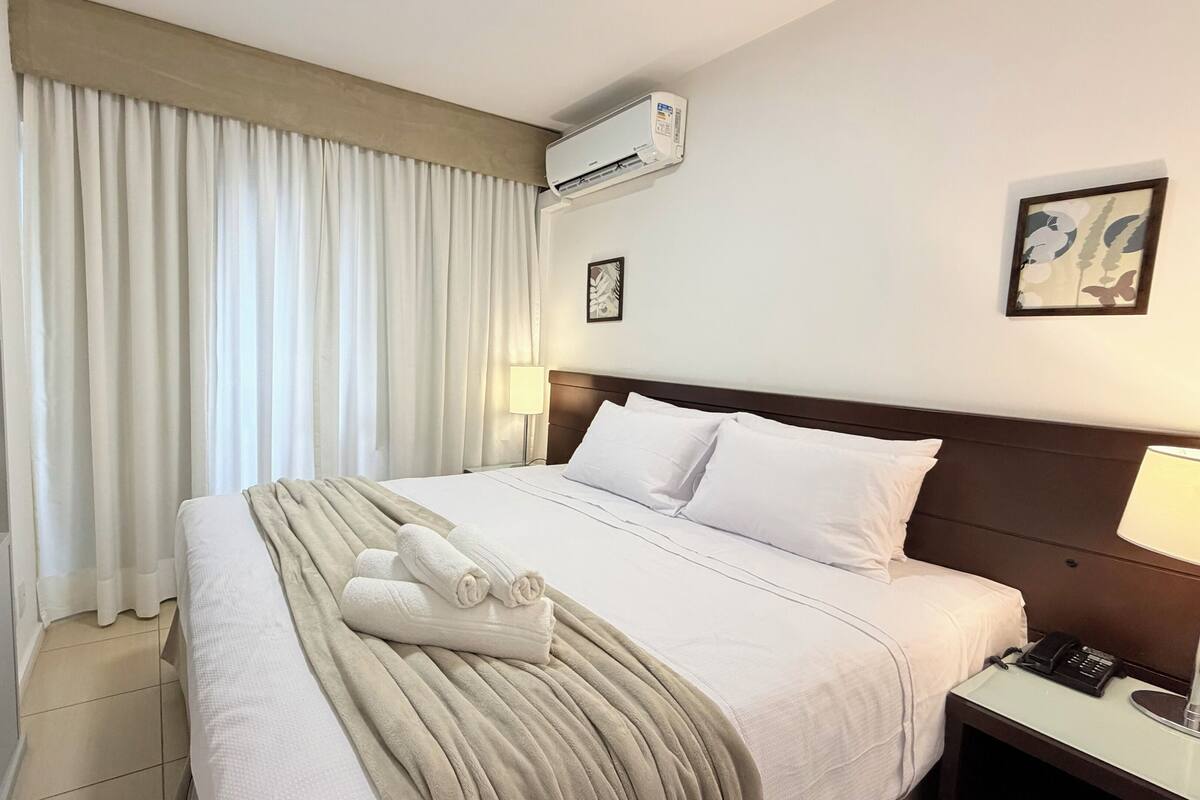 Une chambre d’hôtel avec un lit bien fait, une lampe de chevet, un téléviseur posé sur un meuble et un appareil de climatisation fixé au mur.