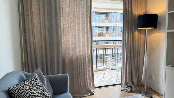 Un salon moderne avec un canapé gris, un coussin à motifs et une lampe sur pied. On aperçoit, à travers les rideaux, un balcon avec une chaise blanche et une balustrade.