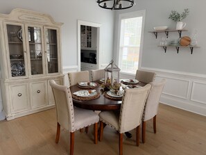 A dining room with a round table set for six, surrounded by six upholstered chairs with button details. A white cabinet with glass doors is to the left, displaying silverware and dishes. - 2026 Masters Week Rental (Grovetown)
