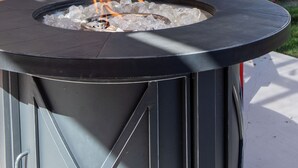 Un foyer extérieur de forme ronde, avec une structure en métal noir, entouré d’une clôture en bois et agrémenté d’un banc rouge.