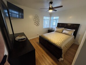 Ein Schlafzimmer mit einem Bett, einem an der Wand montierten Fernseher, einem Deckenventilator und einem Fenster mit Jalousien.