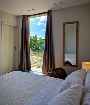 Une chambre à coucher avec un grand lit, un miroir et une vue sur un lac et des arbres, visible à travers des rideaux ouverts.