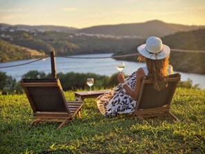 Une personne assise sur une chaise, avec un ordinateur portable et un verre de vin, et qui contemple un paysage magnifique sur un lac et des collines.