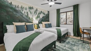 Ein Hotelzimmer mit zwei Betten, einem Deckenventilator, einem Fenster mit Vorhängen und einem Waldgemälde an der Wand.
