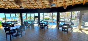 Ein Restaurant mit großen Fenstern, die einen Pool und den Ozean zeigen. Tische und Stühle sind im Inneren aufgestellt.