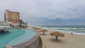 A beachside pool with a stone wall, thatched umbrellas, and a hotel building in the background. - Baja 404 Las Palmas Beachfront Resort (Puerto Peñasco)