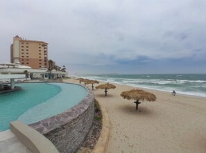Ein Pool am Strand mit einer Steinmauer, reetgedeckten Sonnenschirmen und einem Hotel im Hintergrund.