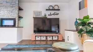Een woonkamer met een stenen muur, een flatscreen televisie op een roze standaard, een boekenkast en een grote groene plant.