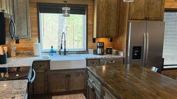 A kitchen with wooden cabinets, a stainless steel refrigerator, a granite countertop, and a window with a view of trees.