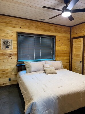 Une chambre à coucher avec un lit, un ventilateur de plafond, une fenêtre avec des stores et un mur en bois orné d’une inscription.