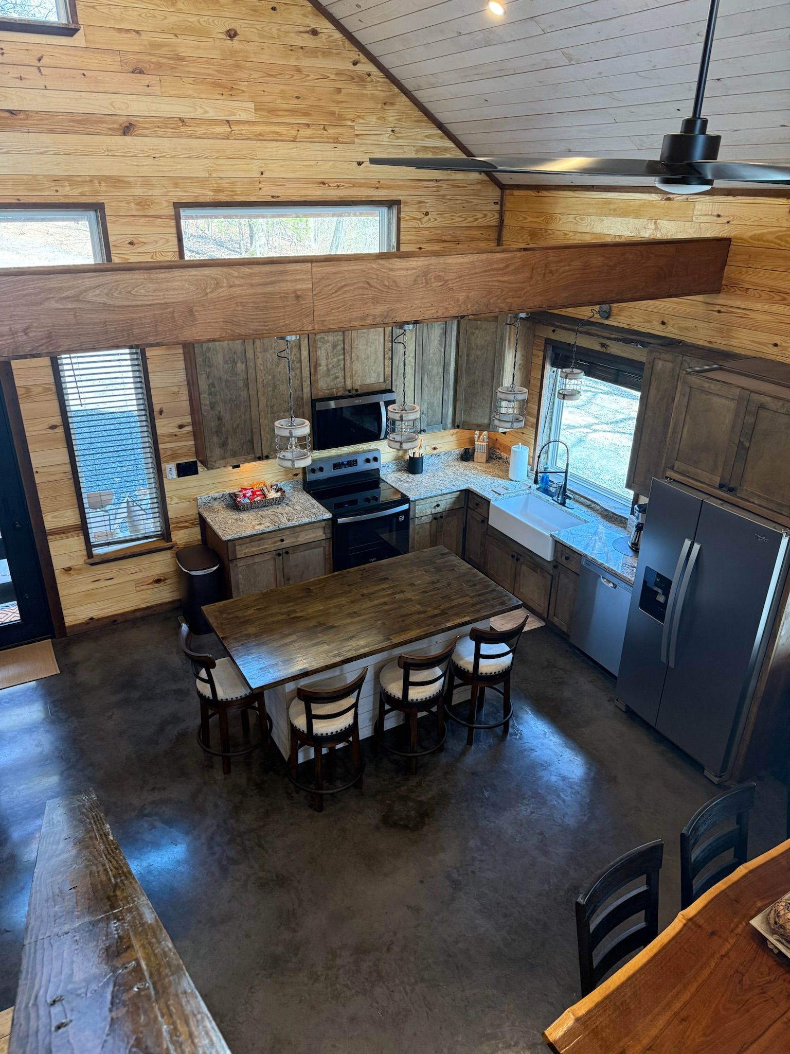A high-ceilinged kitchen with wooden walls, a central island with stools, a refrigerator, and a microwave.